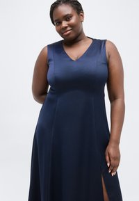 Γυναίκα με πλεξούδες, που φοράει αμάνικο μπλε φόρεμα με ανοίγματα στο πλάι, χαμογελώντας απαλά μπροστά σε απλό φωτεινό φόντο.