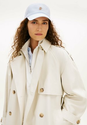 Frau mit lockigem Haar, die einen hellbeigen Trenchcoat, ein hellblaues Oberteil mit Reißverschluss und eine hellblaue Kappe mit kleinem goldenem Emblem trägt.