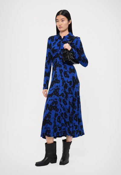 Plava i crna haljina s uzorkom, dugim rukavima i ovratnikom, s naboranim suknjom. Također su vidljive crne kožne čizme i mala torbica.