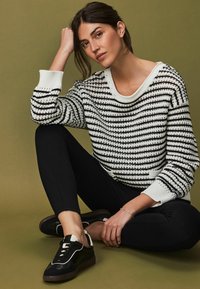 Zwart-wit gestreepte gebreide trui met een brede halslijn, zwarte legging en zwarte sneakers met witte accenten, tegen een groene achtergrond.