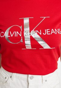 Červené tričko Calvin Klein s velkým stříbrným logem "CK" a bílým textem "CALVIN KLEIN JEANS", spárované s bílými kalhotami s vysokým pasem.