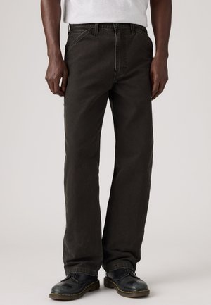 Mann, der schwarze Jeans mit geradem Bein, schwarze Lederschuhe und ein weißes, in die Hose gestecktes Hemd trägt, steht vor einem einfarbigen Hintergrund.