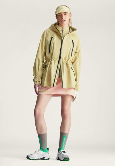 Chaqueta impermeable amarilla con cremalleras negras y cintura ajustable, combinada con una falda rosa con detalles en verde y calcetas grises con zapatos verdes.
