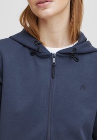 Felpa con zip di colore blu navy, dotata di cappuccio con coulisse, con una texture liscia e dettagli neri. Logo discreto sul petto.