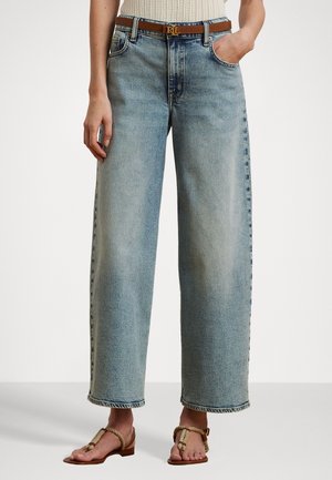 Vrouw draagt hoge taille, vervaagde blauwe jeans met wijde pijpen, een bruine leren riem met gouden gesp, een beige gebreide top en beige sandalen met koordriempjes.