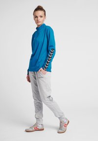 Maglietta sportiva a maniche lunghe turchese con colletto alto e motivo a chevron nero sulle maniche, abbinata a pantaloni felpati grigi chiari con logo.