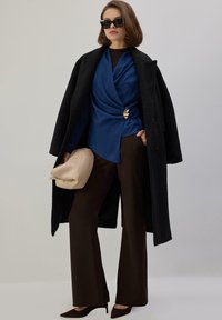Abrigo negro, blusa envolvente de satén azul con broche dorado, pantalones marrones de pierna ancha, bolso de mano beige y tacones negros puntiagudos. Gafas de sol completan el look.
