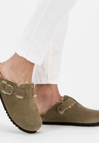 Sabots en daim marron avec des accents en fourrure, sangle à boucle réglable et semelle noire texturée, portés avec un jean blanc.
