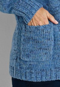 Hand in der Tasche eines blauen Strickpullovers mit melierter Textur, sichtbarem Bündchen und geripptem Saum, vor einem schlichten grauen Hintergrund.