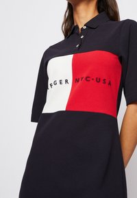 Polo tričko v námořnické modři s výrazným blokovým designem v červené, bílé a modré barvě. Vyšité text "TOMMY HILFIGER NYC USA" v černé na přední straně.