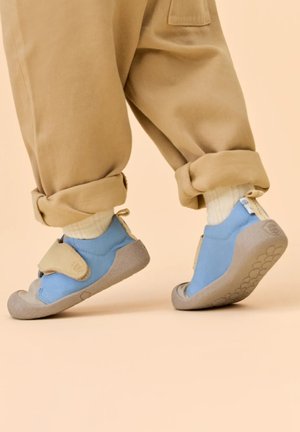 Bambino che indossa pantaloni beige e scarpe morbide blu con strappi in Velcro, sollevando un piede su uno sfondo neutro.
