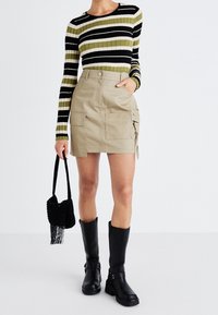 Pruhovaný černý, zelený a bílý žebrovaný top s dlouhým rukávem, spárovaný s béžovou cargo sukní se dvěma bočními kapsami, nošený s černými kozačkami přes kolena.