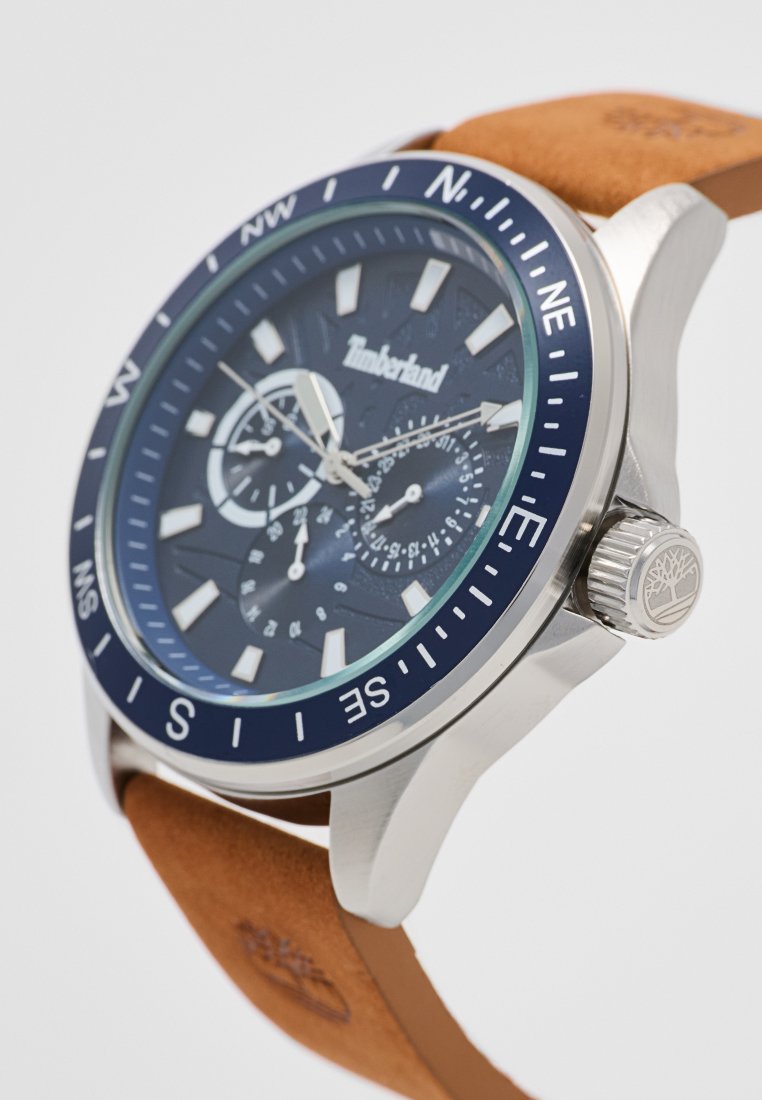 Reloj de pulsera analógico con esfera azul marino, caja de acero inoxidable plateado, correa de cuero marrón, marcadores luminosos y marcas de brújula en el bisel.