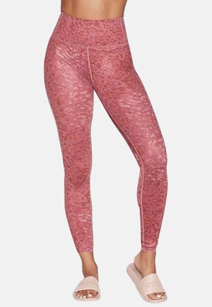 Woman wearing high-waist pink patterned leggings and light pink slide sandals, standing with legs slightly apart on a white background.