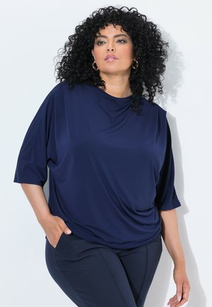 Donna con capelli neri ricci che indossa una maglia larga blu navy e pantaloni scuri, in piedi con una mano in tasca su sfondo bianco.