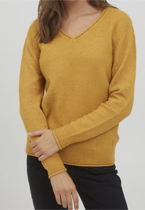 Kvinde iført sennepsgul V-hals sweater med ribstrikkede manchetter og kant, kombineret med sorte bukser, armene krydset foran.