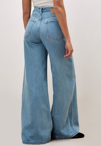 Jeans large de couleur bleu clair, taille haute, avec deux poches arrière et une patch en cuir sur la taille. Texture denim lisse.