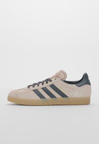 Béžové semišové tenisky Adidas Gazelle s černými pruhy, černými tkaničkami, béžovým jazykem a gumovou podrážkou na bílém pozadí.