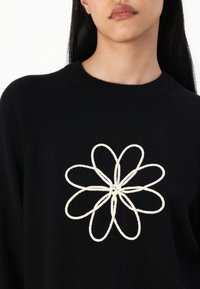 Svart genser med et stort hvitt brodert blomsterdesign i midten, rund halsutskjæring og myk tekstur.
