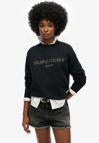 Marineblauwe sweatshirt met "SUPERDRY PARIS" in rhinestones, gedragen over een witte overhemd, gecombineerd met zwarte denim shorts met rafelige randen.
