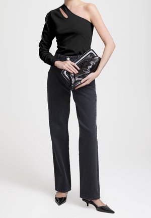 Vrouw draagt een zwarte asymmetrische top, zwarte jeans met hoge taille, zwarte puntige hakken en houdt een zwarte clutch vast.