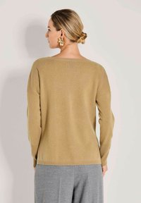 Frau mit blonden Haaren in einem tiefen Dutt, die einen beigen Pullover und graue Hosen trägt, von hinten blickend, vor einem schlichten hellen Hintergrund.