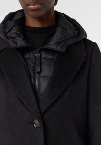 Cappotto nero con un leggero cappuccio imbottito nero. Il cappotto presenta una chiusura a un bottone e un tessuto texturizzato, che offre calore e stile.