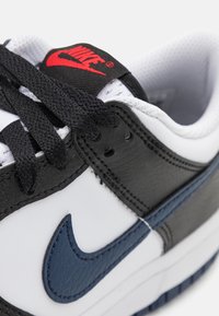 Baltos sportiniai batai su juodais odiniais akcentais, tamsiai mėlyna detalizacija, juodomis raištelėmis ir raudonu Nike logotipu ant liežuvėlio. Lygūs ir tekstūruoti paviršiai.