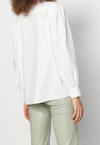 Camicia bianca a maniche lunghe con spalle a sbuffo, dotata di polsini con bottoni e orlo dritto; abbinata a pantaloni aderenti di un verde chiaro.
