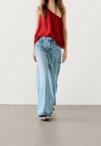 Haut rouge à une épaule, fluide, associé à un jean en denim bleu clair à jambe large. Le mannequin porte des sandales plates noires en marchant sur un sol lisse.
