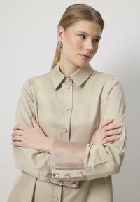 Chemise beige en soie à boutons avec des coutures pressées, un col classique et un tissu transparent avec des poignets ornés. Texture lisse et design ajusté.