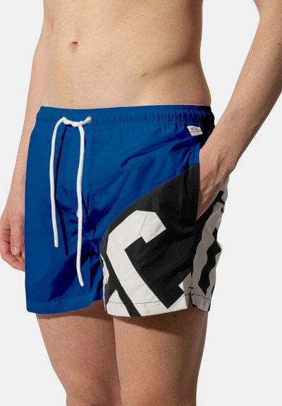 Uomo che indossa pantaloncini da bagno blu con coulisse bianca e motivo geometrico nero e bianco sul lato destro, mano in tasca.