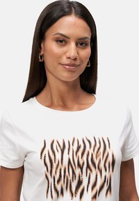 T-shirt blanc présentant un design de motif animal stylisé et rayé en noir et orange avec des embellissements. Encolure ronde et manches courtes.