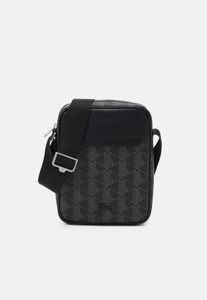 Sac bandoulière noir avec sangle ajustable, motif géométrique, logo Lacoste embossé et compartiment principal zippé.