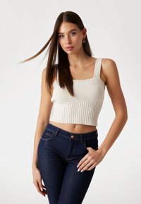 Gebreid cropped top in crème met ribbeltextuur en brede banden, gecombineerd met donkerblauwe slim fit jeans met een knoop en voorkennis.