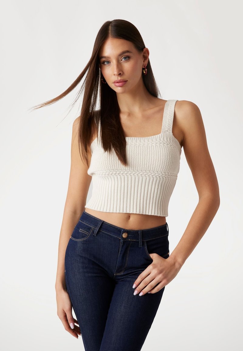 Gebreid cropped top in crème met ribbeltextuur en brede banden, gecombineerd met donkerblauwe slim fit jeans met een knoop en voorkennis.