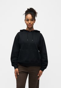 Jeune femme aux cheveux bouclés portant un sweat à capuche noir et un pantalon marron, debout droite devant un fond blanc uni.