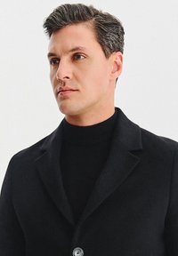 Czarny płaszcz z kołnierzem i zapięciem na jeden guzik, noszony na czarnym golfie. Gładka tekstura, nowoczesny design, dopasowana sylwetka.