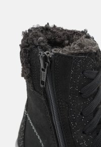 Botín de ante negro hasta el tobillo con un cuello de piel texturizada, cierre de cremallera y cordones negros. Presenta sutiles destellos plateados en el exterior.