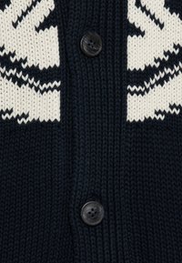 Cardigan en maille bleu marine avec un motif géométrique blanc, quatre boutons noirs et un tissu texturé à côtes.