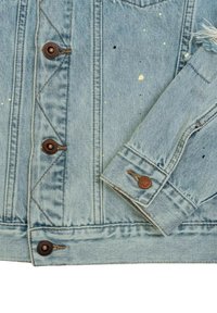 Giubbotto in denim azzurro chiaro con dettagli sfrangiati, bottoni marroni e schizzi di vernice gialla e nera. Presenta un colletto classico e tasche sul petto.