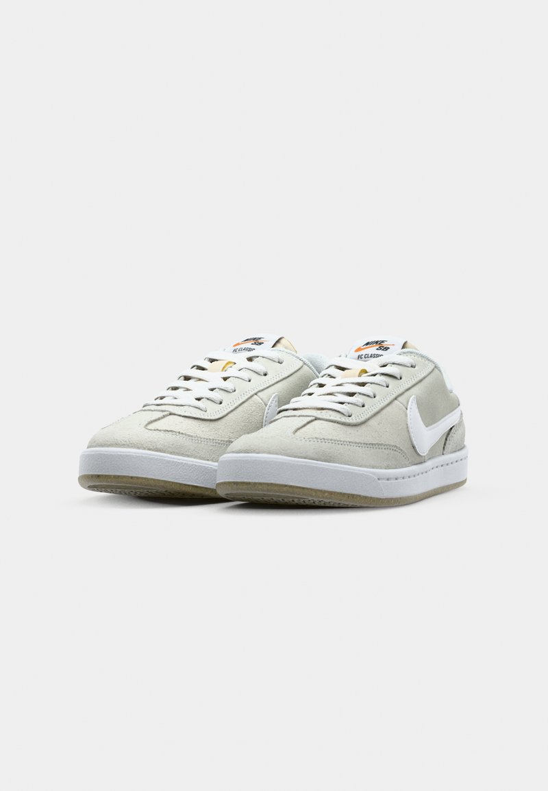 Zapatillas de ante gris claro con acentos en piel blanca y suela blanca. Presentan cordones planos y logotipo en la lengüeta y el costado. Superficie texturizada.