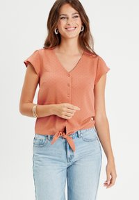 Blouse à manches courtes couleur rouille avec un col en V, détails de boutons sur le devant et un ourlet noué. Tissu doux et texturé ; coupe décontractée.