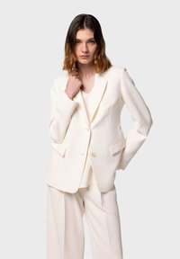 Blazer crema con rever a punta, chiusura a un bottone e due tasche anteriori, abbinato a un top a scollo a V e pantaloni a gamba larga.