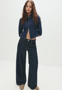 Young woman with long curly hair wears dark blue zip-up denim jacket, wide-leg matching jeans, and beige flats with eyelet details.