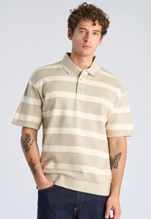 Ung mand med krøllet hår iført beige og hvidstribet kortærmet polotrøje og mørke jeans, der viser tatoveringer på begge underarme.