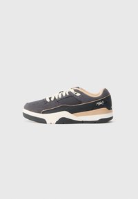 Zapatillas de estilo bajo en gris oscuro y beige con cordones blancos, acentos de suela negra y paneles de tela texturizada sobre un fondo liso.