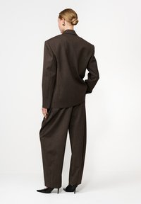 Barna, textúrált anyagú zakó és laza szabású, bőszárú nadrág, fekete, hegyes orrú magassarkú cipővel párosítva. Hátulról nézve.