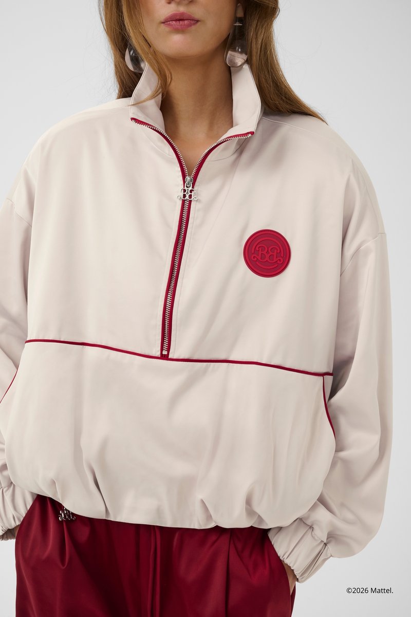 Kvinde iført en lys beige pullover med rød kant, halvlukning og rød logo-patch, kombineret med røde bukser, hænderne i lommerne.
