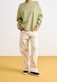 Personne portant un sweat-shirt vert clair avec un texte rose, un pantalon ample crème et des chaussures noires sans lacets, debout sur un sol moutarde.